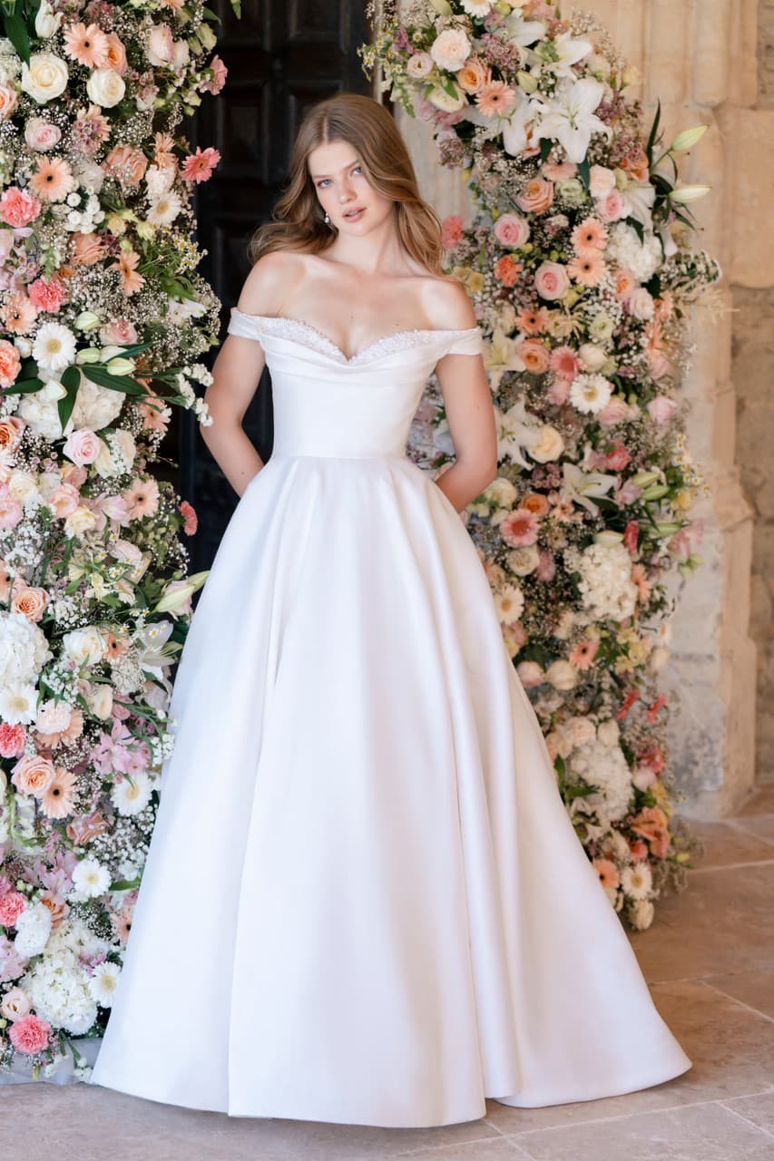 Off-shoulder wedding dress with full skirt standing between floral arrangements